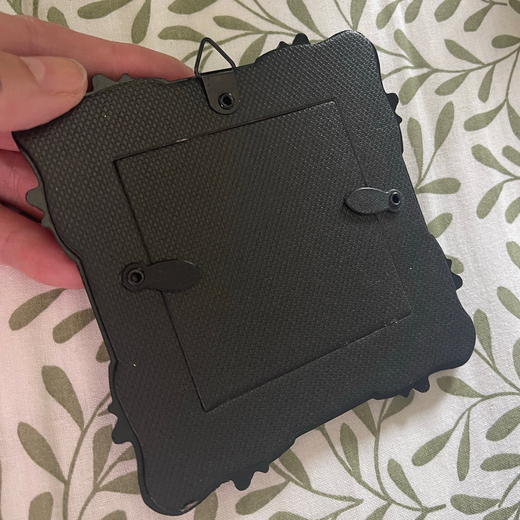 Black textured square picture frame held by a hand on a patterned surface