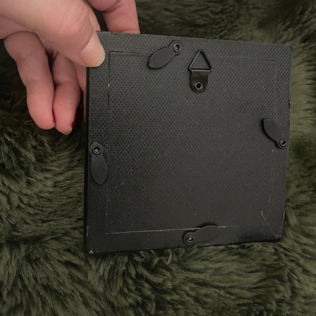 Black rectangular picture frame back  held by a hand on a green textured surface.