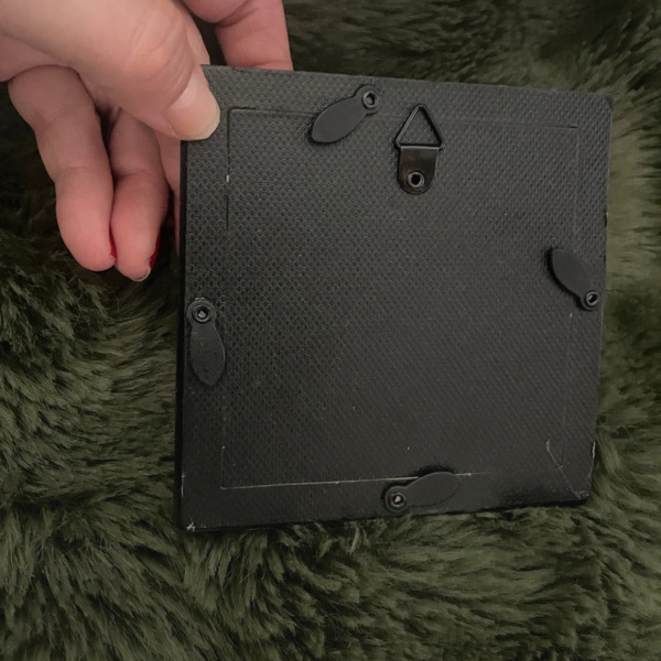 Black rectangular picture frame back  held by a hand on a green textured surface.