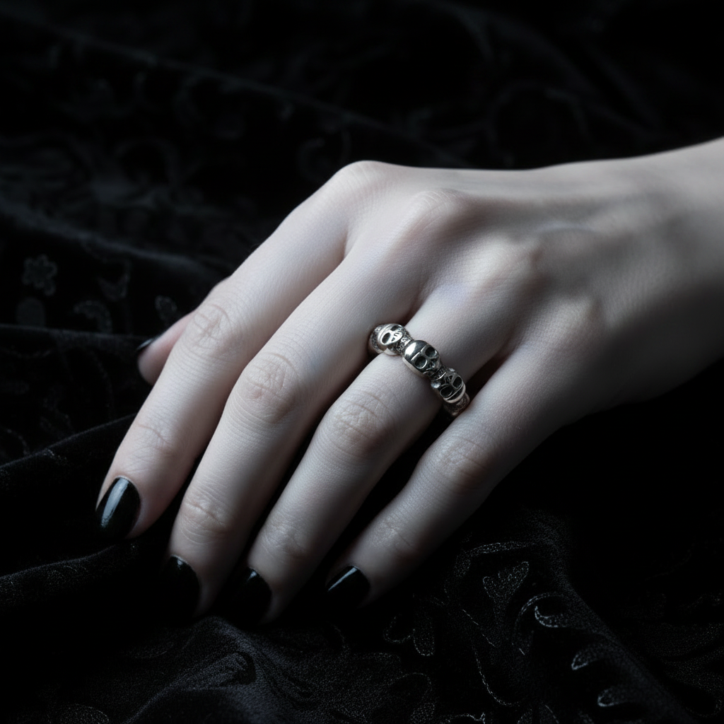 A silver-toned skull ring worn on a hand with black nail polish, resting on a textured black fabric background.