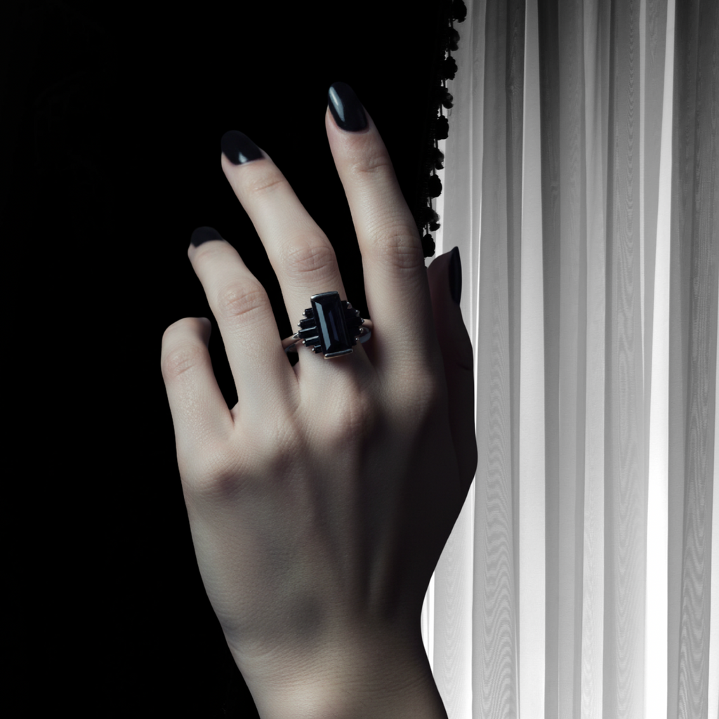 A hand with dark manicured nails wears a black rectangular ring with a silver-toned band, posed against a stark black background and white vertical blinds.