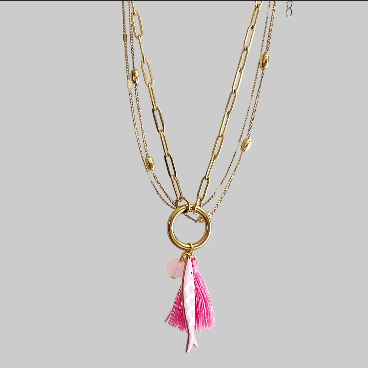 Gold chain necklace with a pink fish, bead and tassel on a grey background