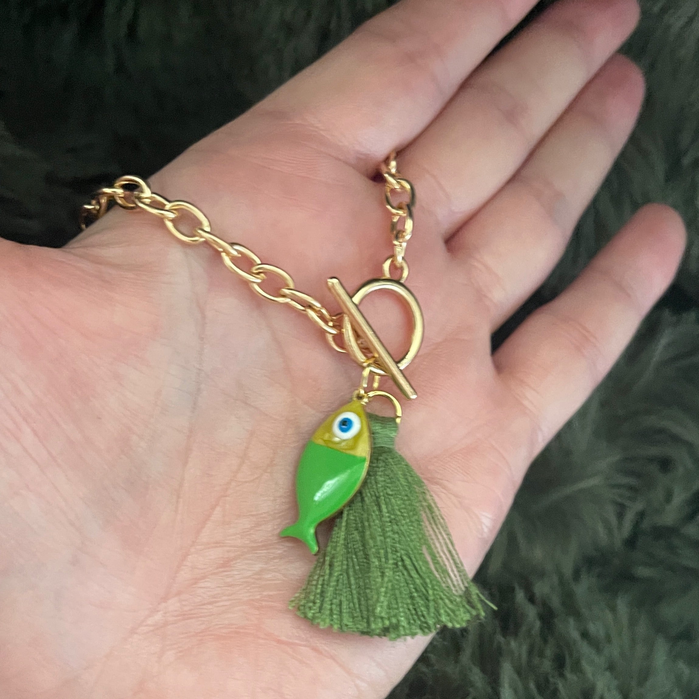 Gold chain bracelet with a green fish charm held in a hand against a dark background