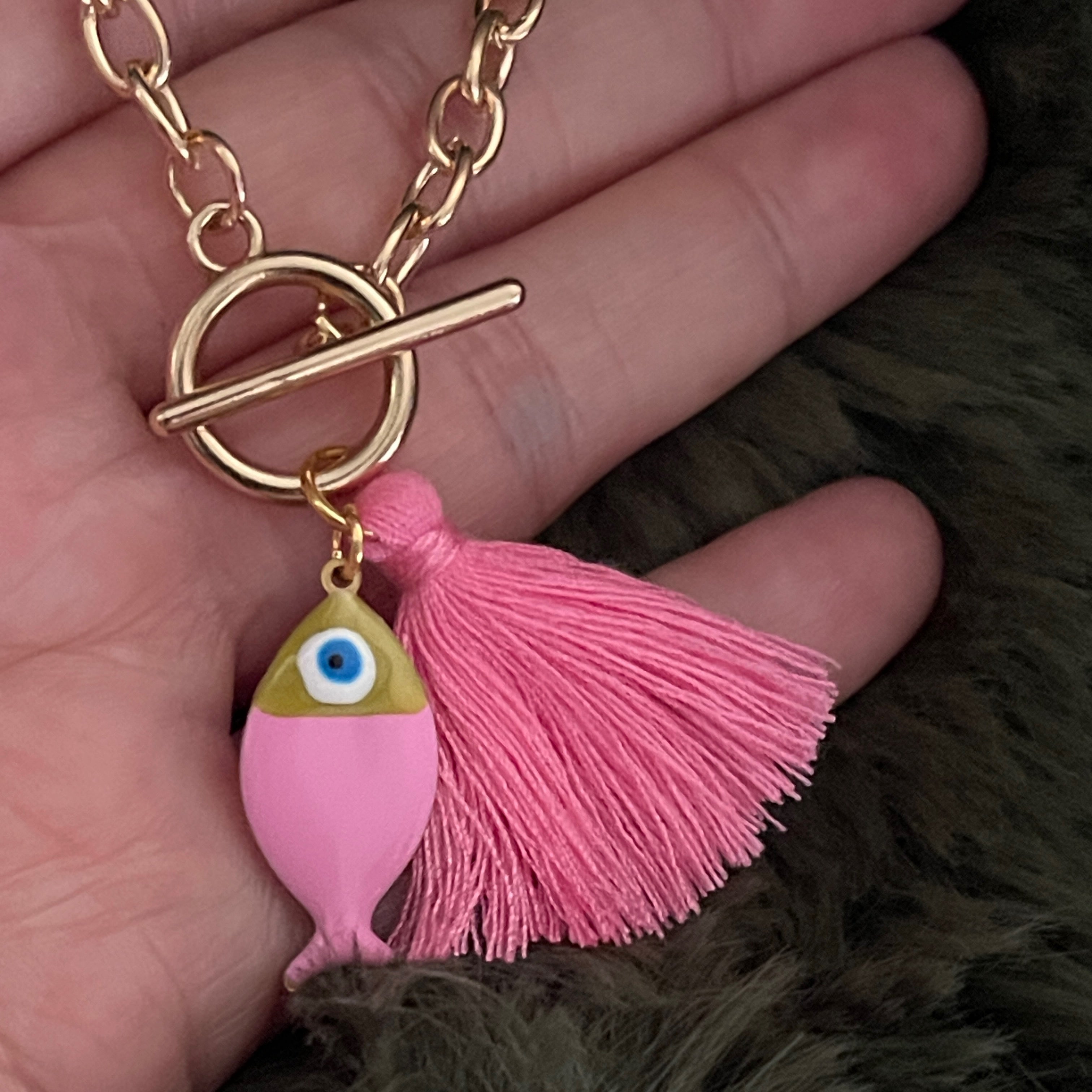 Pink fish charm with tassel on a gold chain held in a hand against a dark background