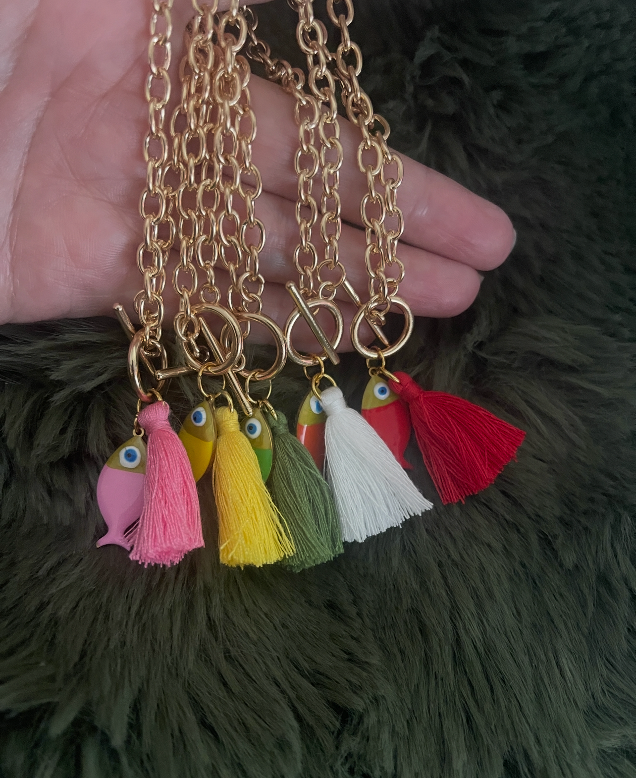 Gold chain necklace with colorful tassel and fish charms held by a hand on a dark textured background