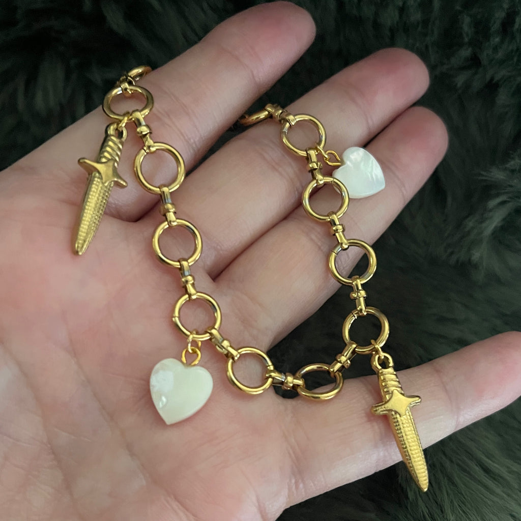 Gold chain bracelet with heart and dagger charms held in a hand against a dark background