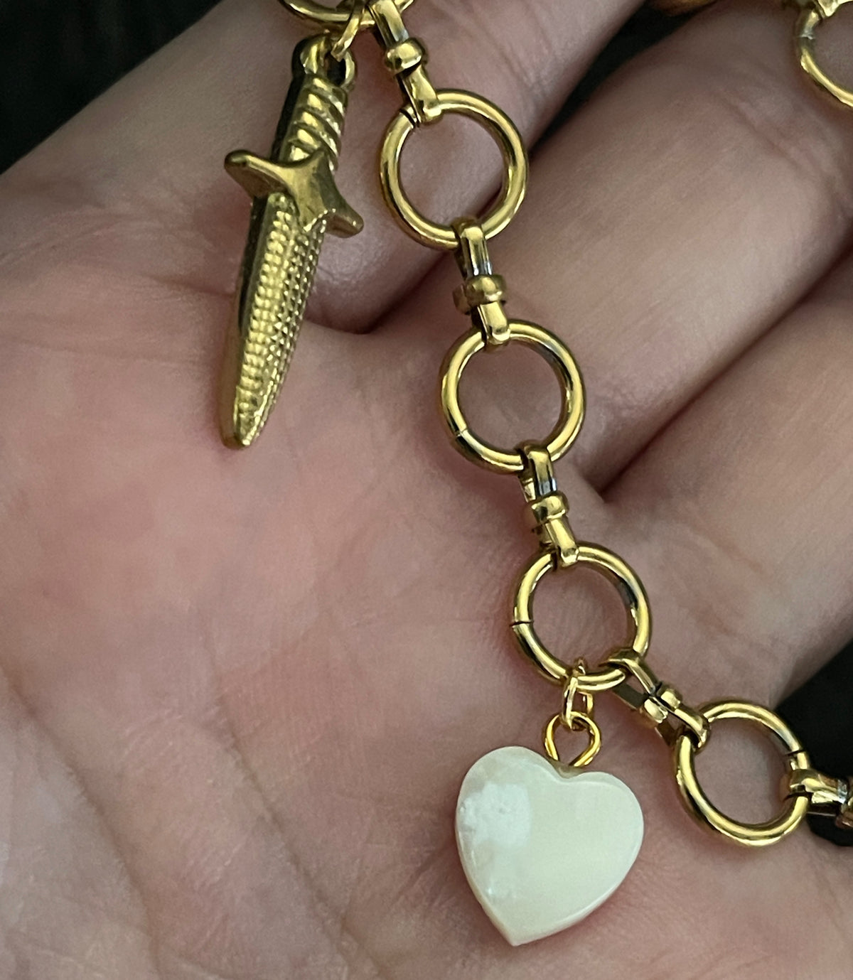 Gold bracelet with a sword and heart charm held in a hand.