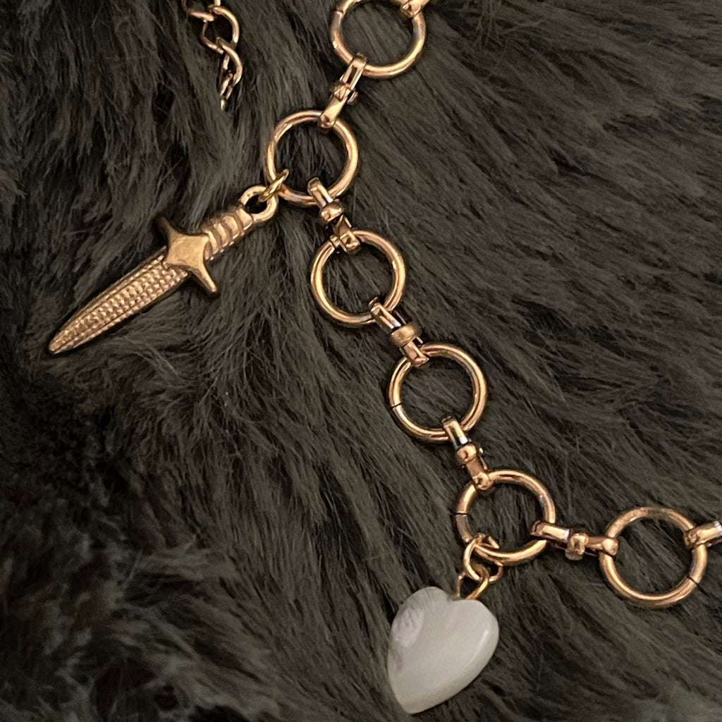Gold chain necklace with dagger and heart charm on a dark fur background