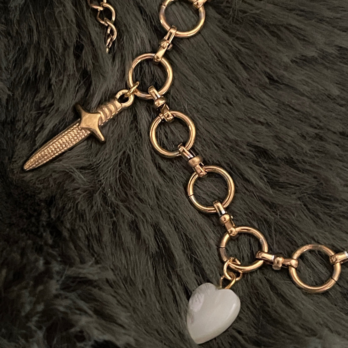 Gold chain necklace with dagger and heart charm on a dark fur background