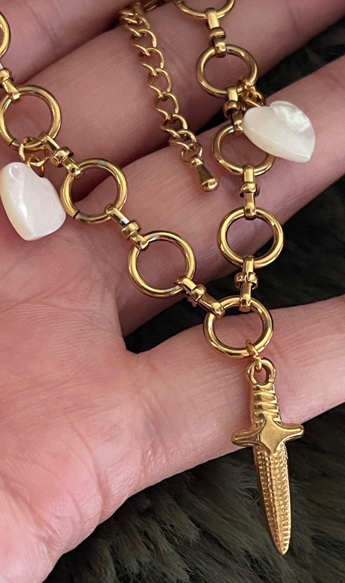 Gold chain bracelet with a dagger charm held in a hand.