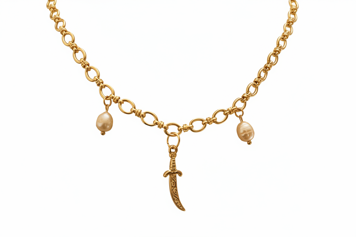Gold-tone chain necklace with a central curved sword charm, flanked by two small oval pearl-like beads on a plain white background.