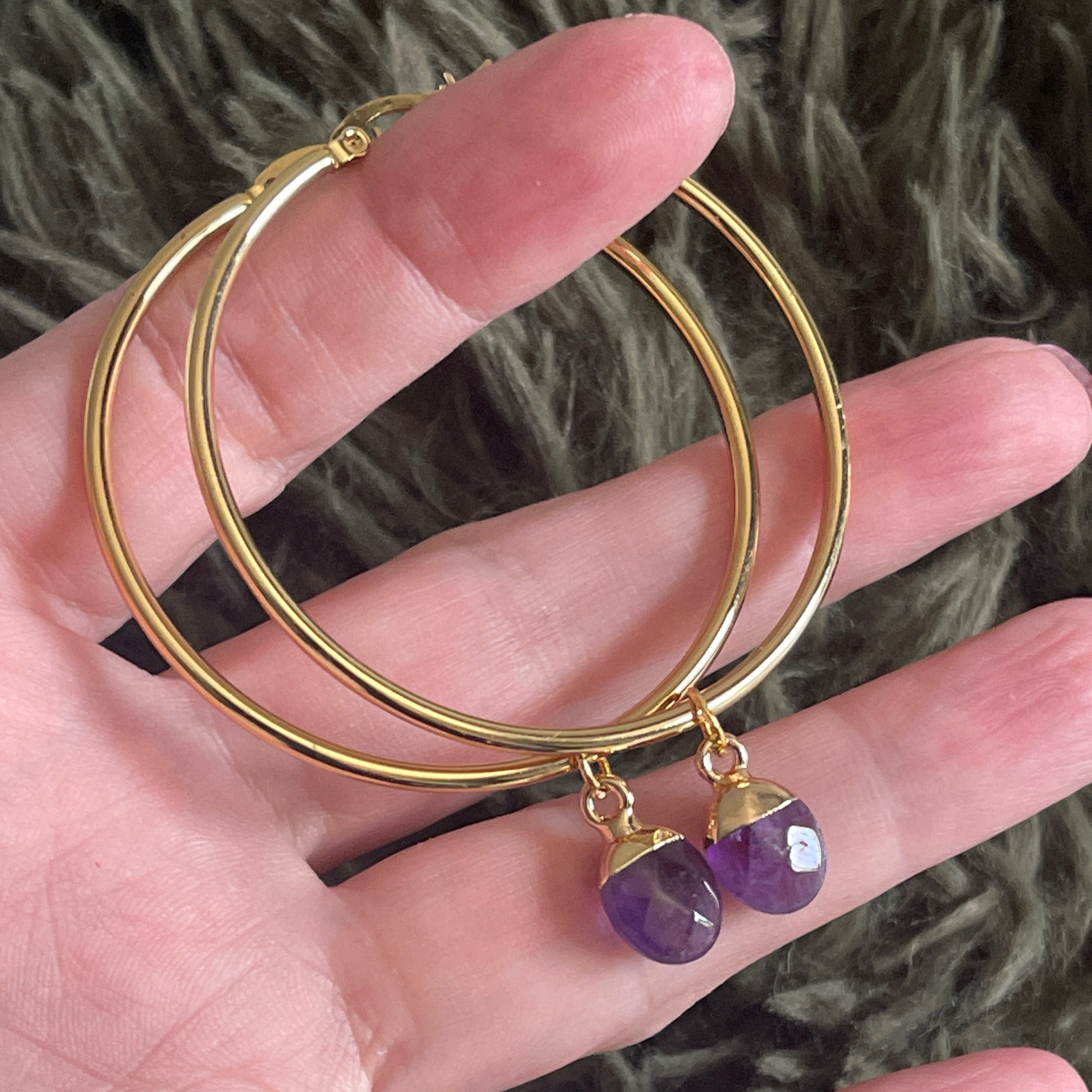 Gold hoop earrings with purple gemstones on a hand against a textured background