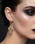 Women with heavy makeup wearing a gold wide cross with ornate scroll detail on it against dark background 