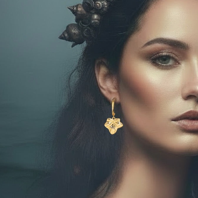 A close-up portrait of a woman wearing a dangling gold earring shaped like a stylized flower or star, with dark hair adorned by a shell-like headpiece against a muted blue-gray background.