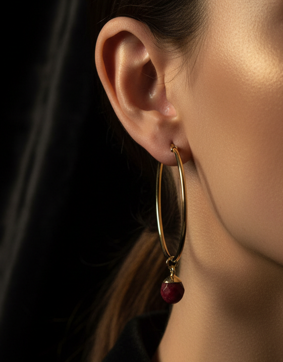 A close-up of a model's ear wearing a gold hoop earring with a dangling faceted dark red gemstone against a dark background.