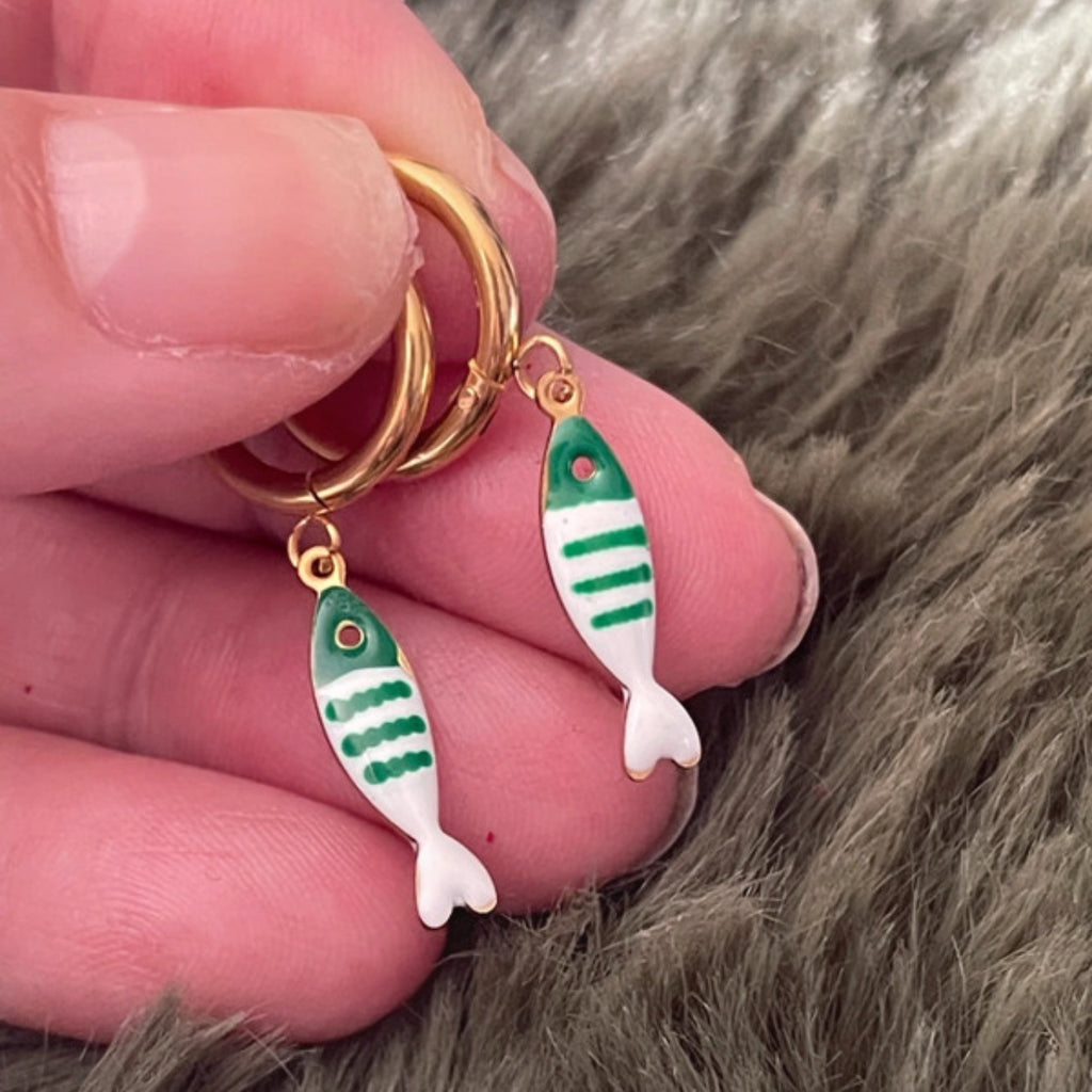 Gold hoop earrings with green and white striped fish charms held between fingers on a fur background.