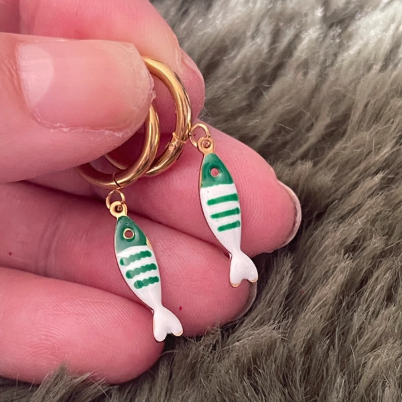 Gold hoop earrings with green and white striped fish charms held between fingers on a fur background.