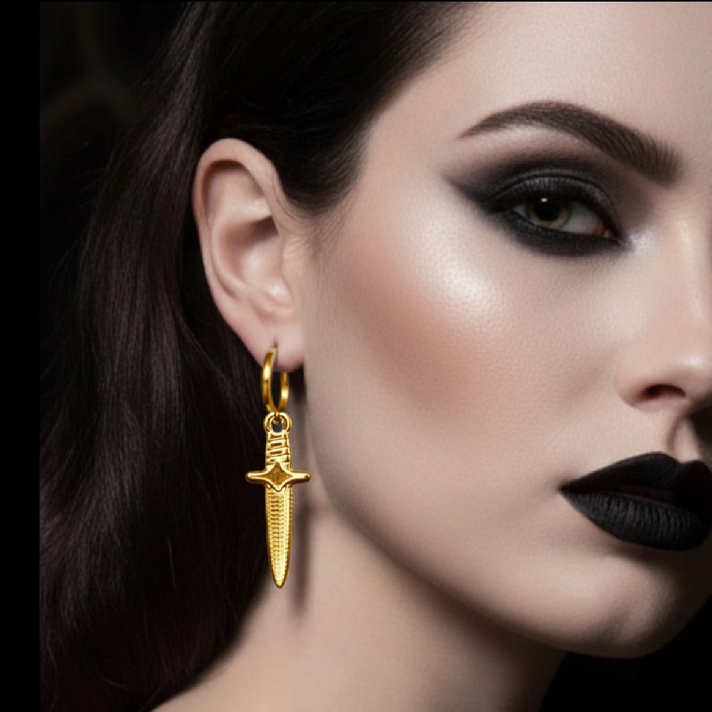 Model with dark makeup wearing a gold hoop earring with a gold dagger a charm against a dark background 
