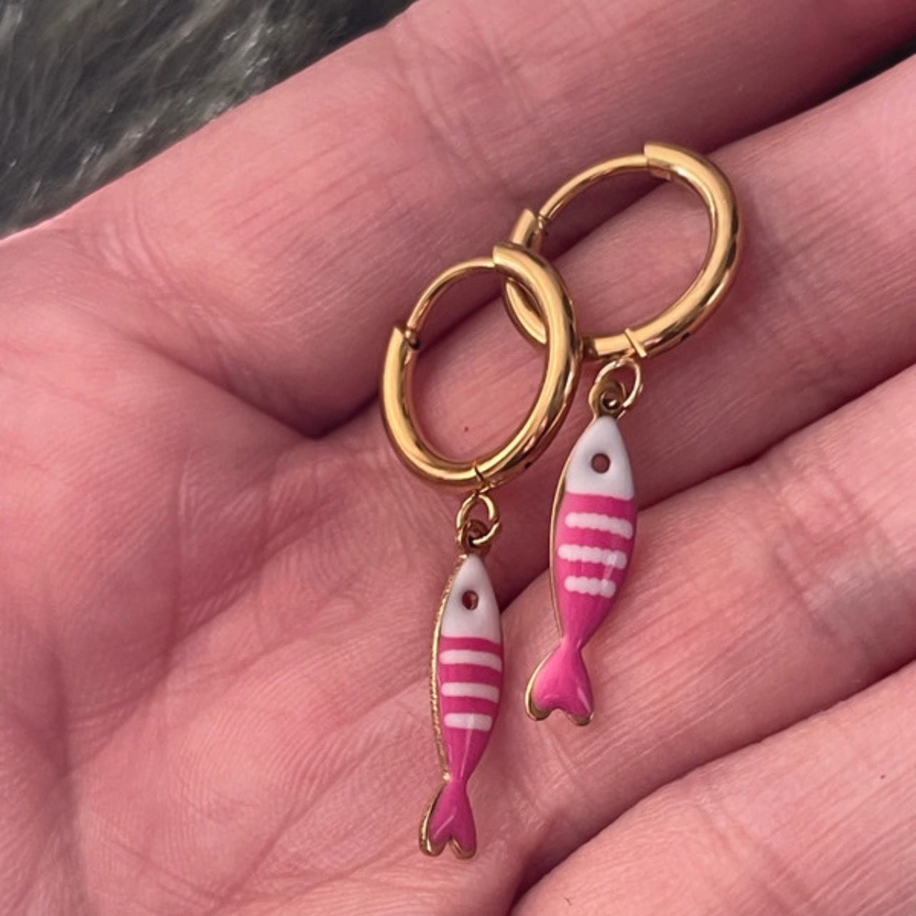 Gold hoop earrings with pink fish charms held in a hand.
