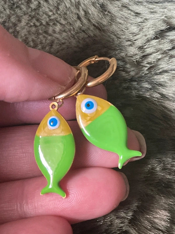 Fish-shaped earrings with gold and green colors held between fingers against a fur background