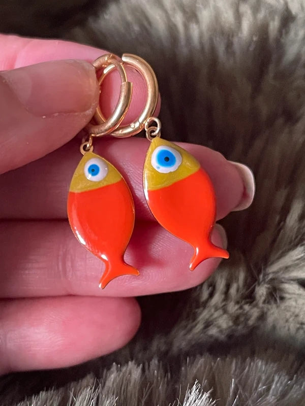 Orange fish-shaped earrings with blue eyes held by a hand against a fur background