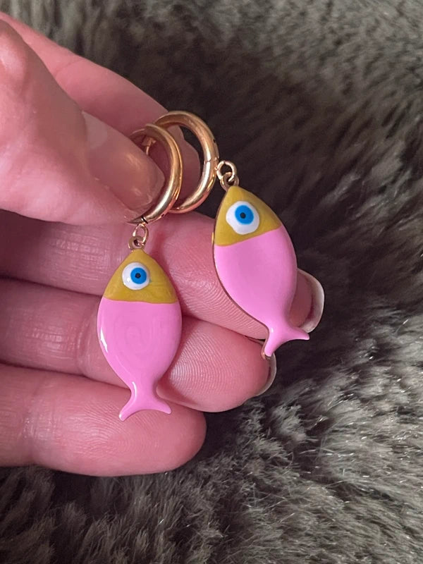 Pink fish-shaped earrings with gold hoops held between fingers against a fur background