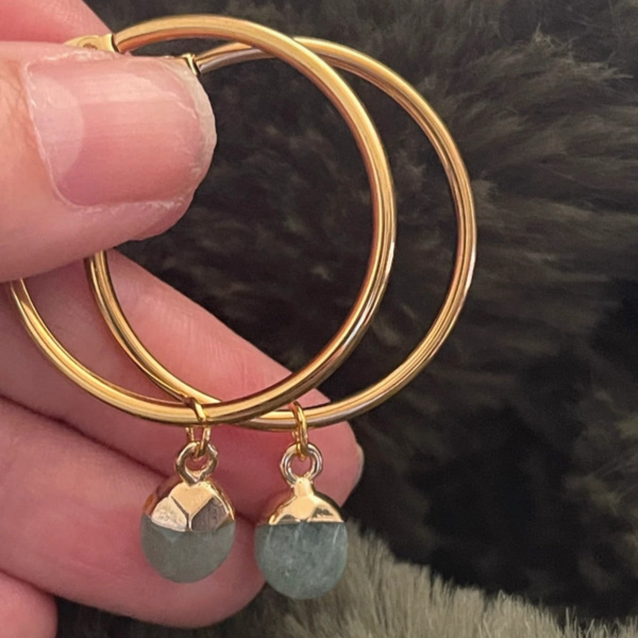 Gold hoop earrings with greenstone drops held by a hand against a dark background