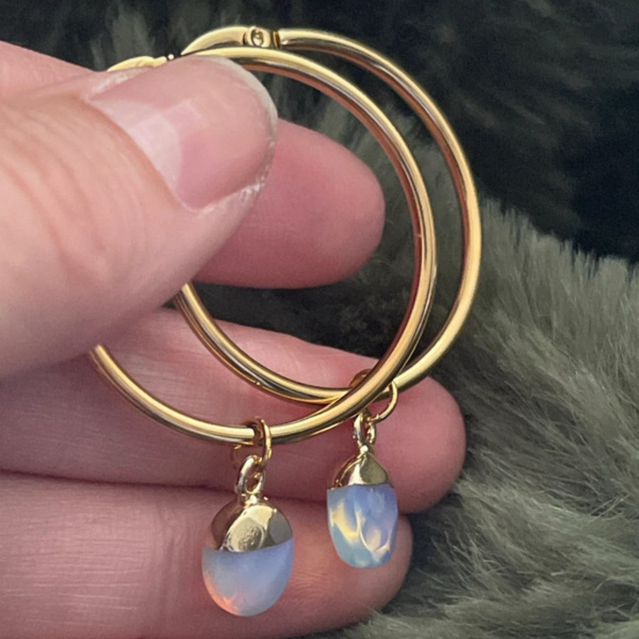 Gold hoop earrings with opalite drops held between fingers against a fur background