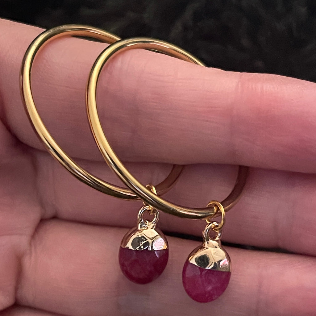 Gold hoop earrings with red gemstone drops held between fingers against a dark background
