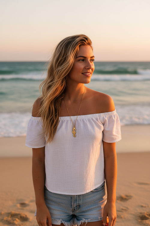 Woman in a white off-shoulder top with a gold seahorse necklace and denim shorts standing on a beach at sunset.