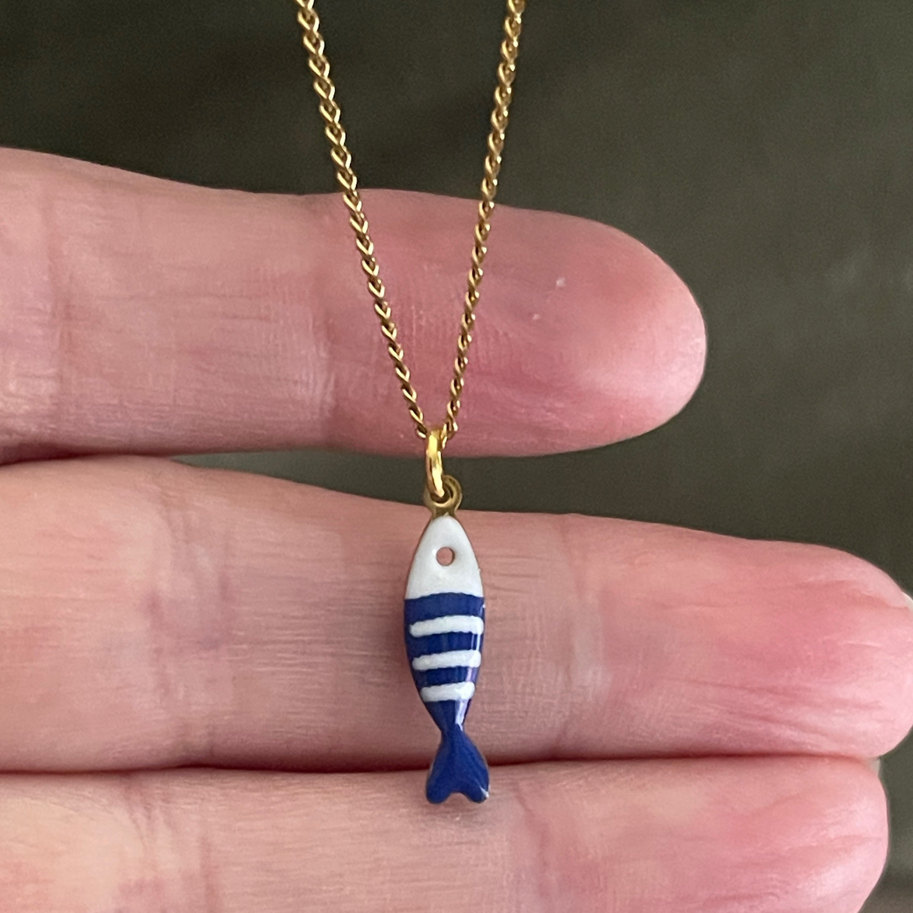 Gold chain with a blue and white striped fish pendant held between fingers against a dark background
