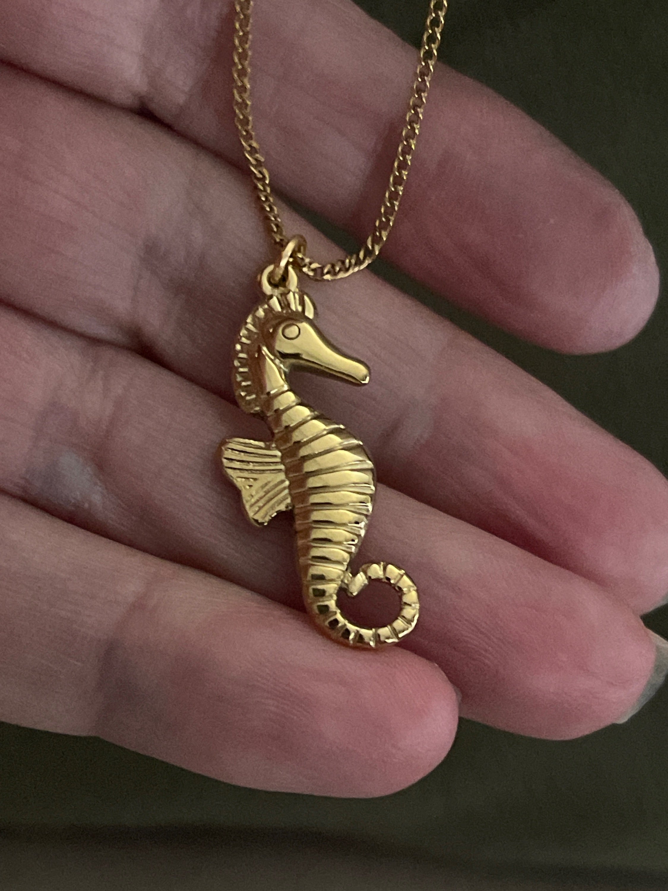 Gold seahorse pendant on a chain held in a hand.