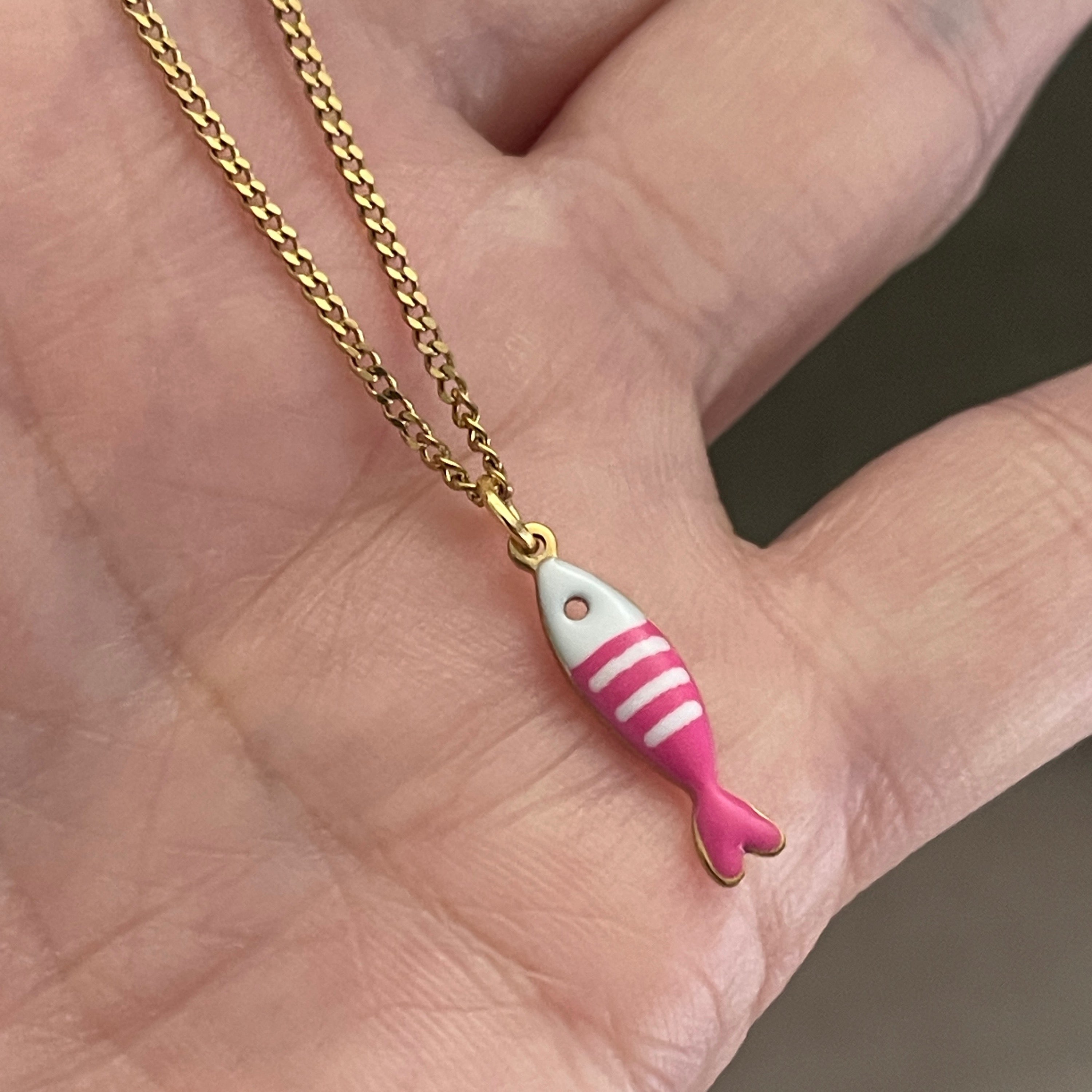 Pink fish-shaped pendant on a gold chain held in a hand.