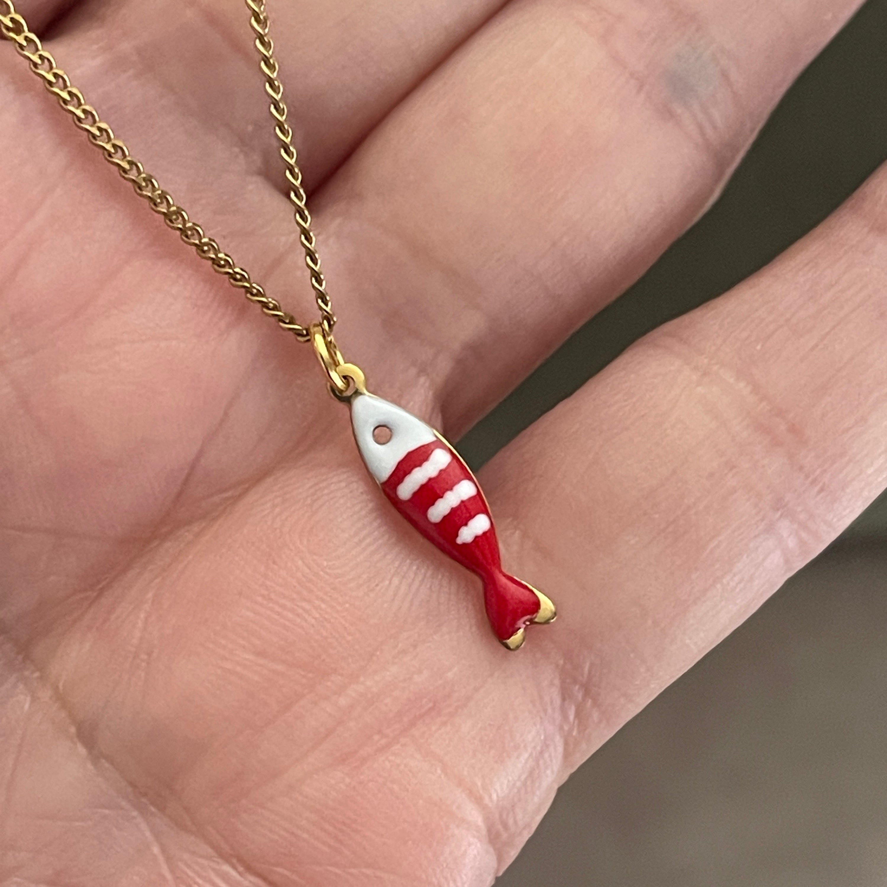 Red and white fish-shaped pendant on a gold chain held between fingers.