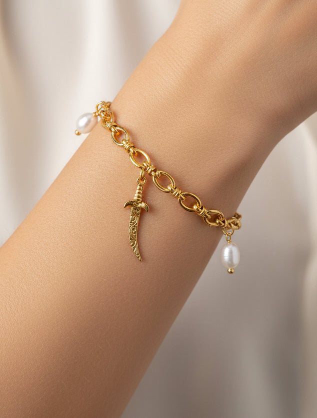 A gold chain bracelet with a dangling sword charm and small pearl beads, shown worn on a person's wrist against a soft light background.