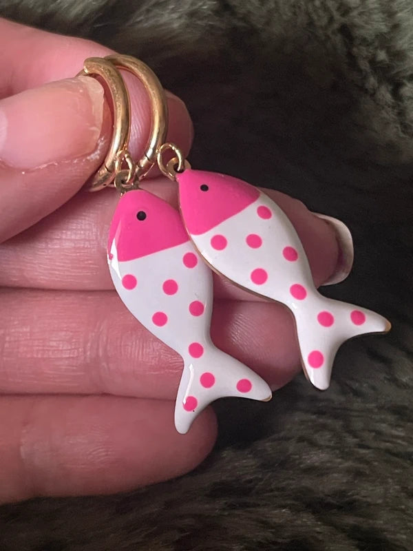Pink polka dot fish-shaped earrings held between fingers against a textured background