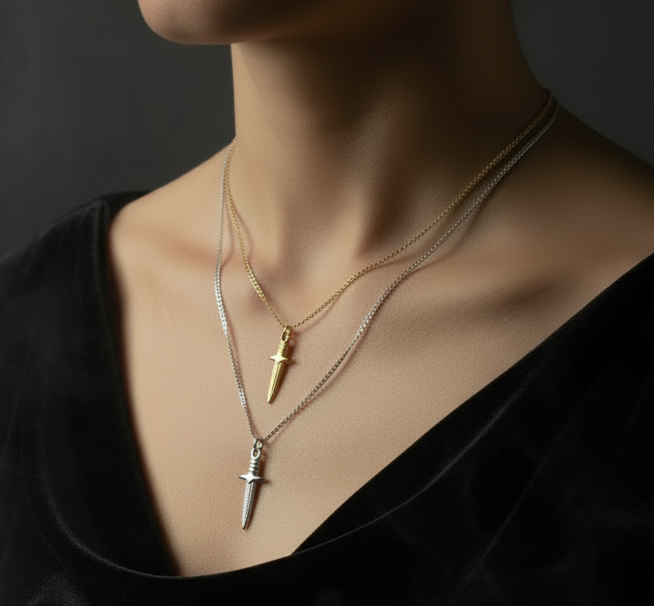 A close-up of a person's neck and chest wearing two layered necklace pendants shaped like swords, one gold and one silver, over a black top against a dark background.