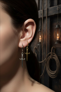 A close-up of a person's ear wearing two dangling earrings: a silver dagger earring and a gold cross earring, set against a dark rustic background with ropes and hanging lanterns.