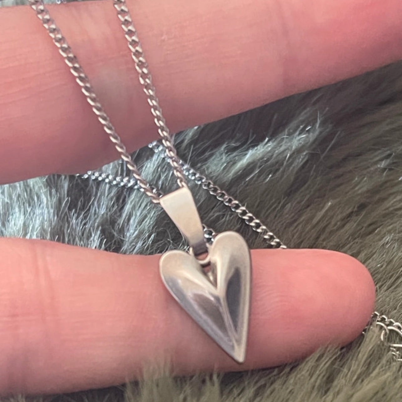 Silver heart-shaped pendant on a chain held between fingers with a blurred background