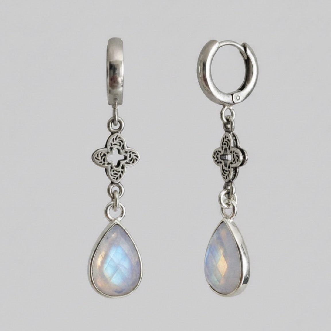 A pair of silver dangle earrings with teardrop-shaped iridescent moonstone drops and ornate filigree details, shown against a solid grey background.