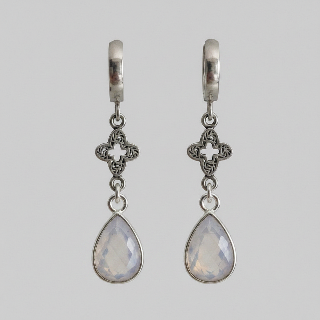 A pair of silver huggie hoop earrings with ornate filigree clover charms and teardrop-shaped milky white gemstone drops, shown against a solid grey background.