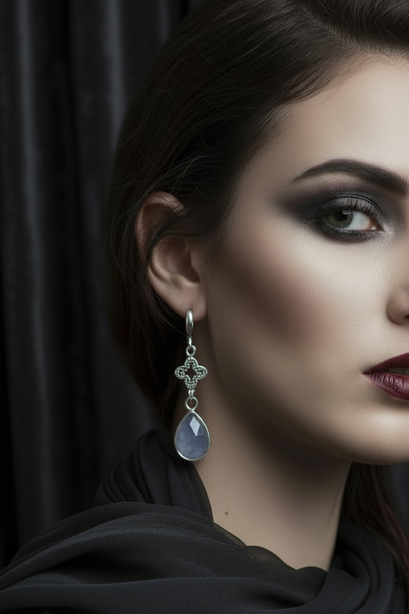 A close-up portrait of a woman wearing a silver-toned drop earring with a small ornate cross-like charm and a teardrop-shaped light blue gemstone, against a dark draped background.