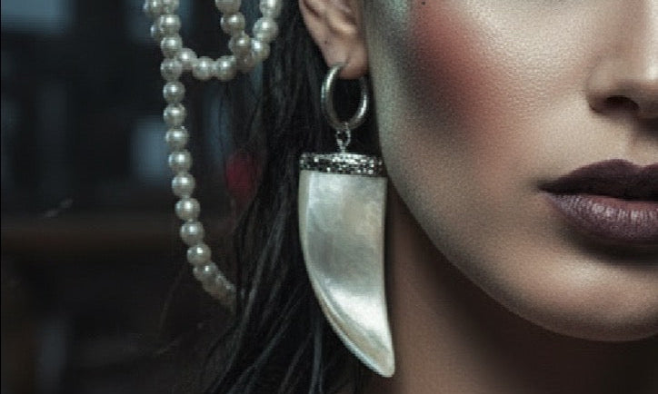 A close-up portrait of a woman wearing a dark brown feathered hat adorned with pearl strands, shell-like beads, and gold-toned embellishments, along with dramatic green eye makeup and a large silver teardrop earring.