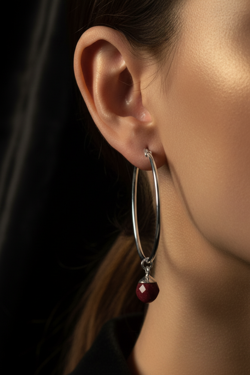 A close-up of a model's ear wearing a silver hoop earring with a dangling faceted dark red bead.