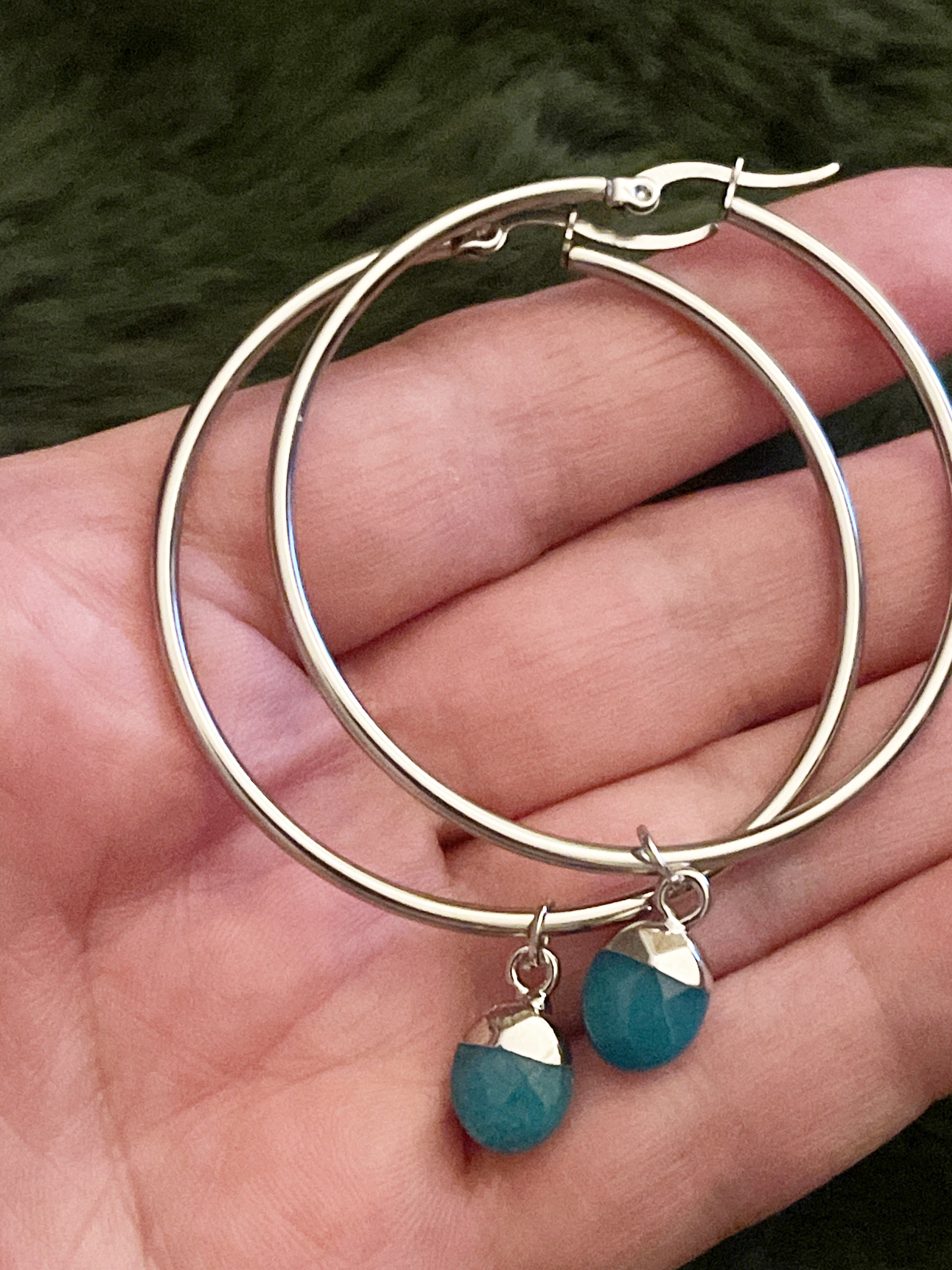 Silver hoop earrings with turquoise crystals held in a hand against a dark background