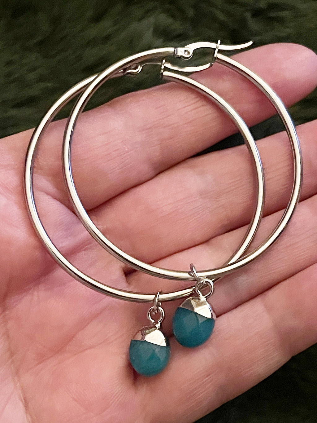 Gold hoop earrings with turquoise crystals held in a hand against a blurred green background
