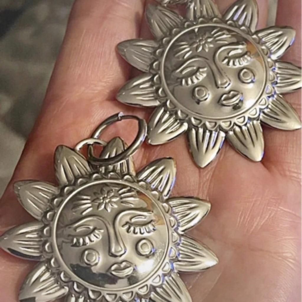 Two silver sun-shaped pendants with face designs held in a hand.