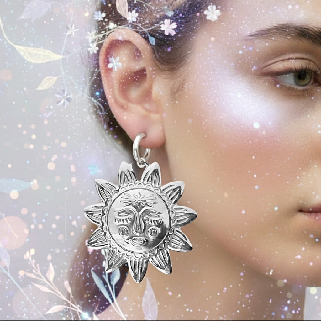 A close-up of a person's ear wearing a dangling silver earring shaped like a sun face, with a soft glittery floral overlay background.