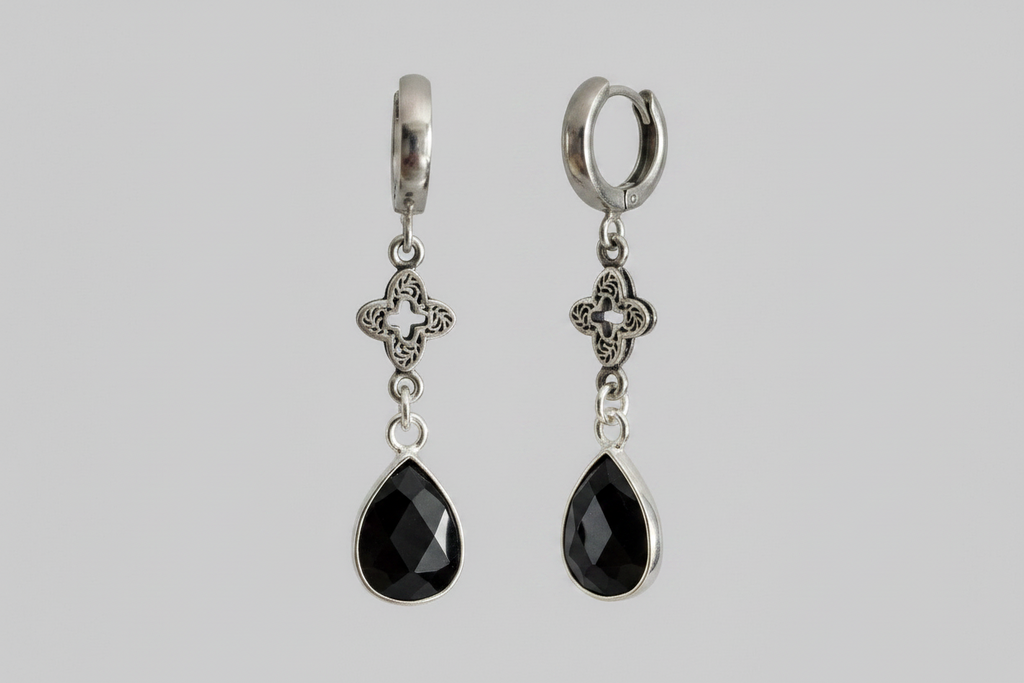 A pair of silver huggie hoop earrings with dangling filigree clover motifs and faceted black teardrop gemstones, shown against a solid gray background.