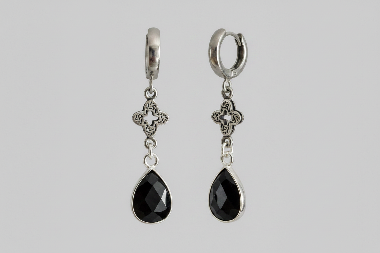 A pair of silver huggie hoop earrings with dangling filigree clover motifs and faceted black teardrop gemstones, shown against a solid gray background.